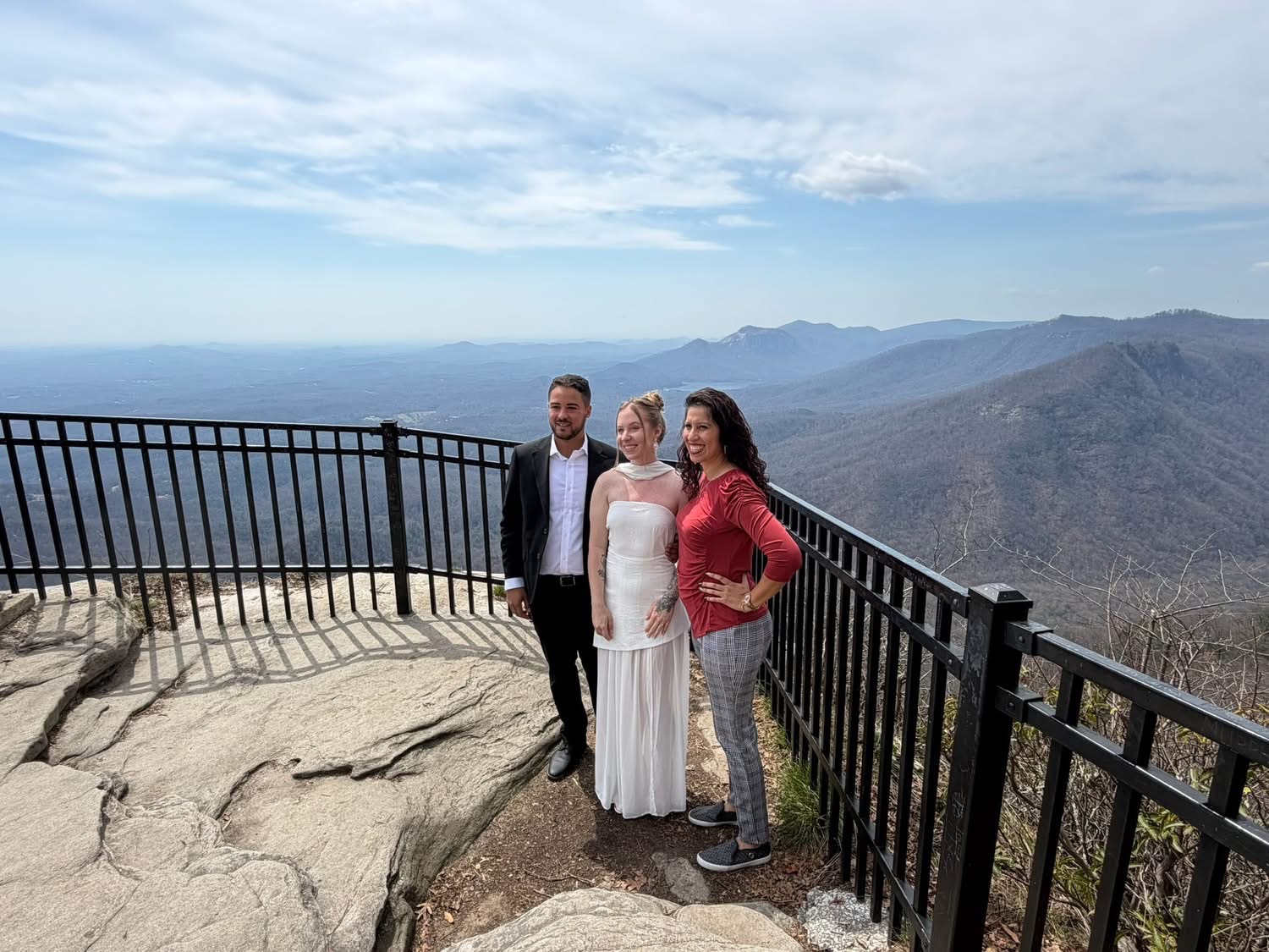 Three people standing on a rocky overlook with a mountain and ocean view in the background, smiling and posing for the photo.