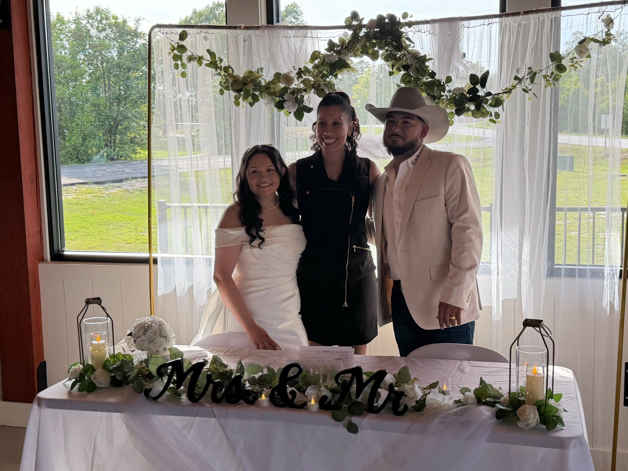 Three people standing behind a decorated table with a wedding sign, in front of a window with greenery outside.