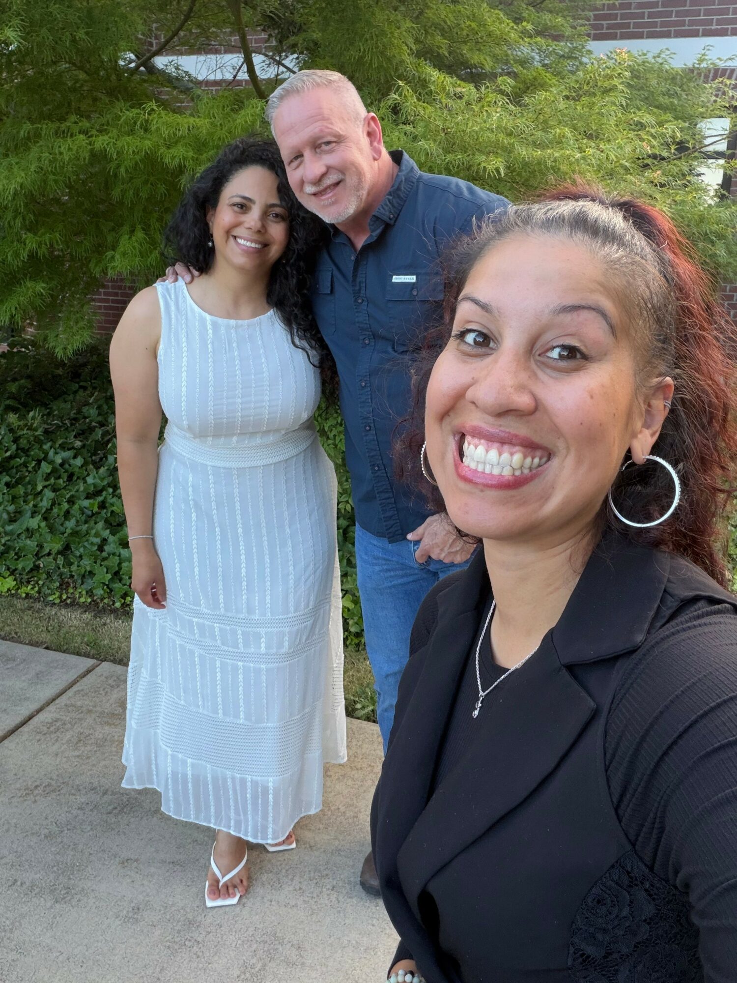 Three people smiling outdoors, woman in black top in foreground, woman in white dress and man in blue shirt in background.