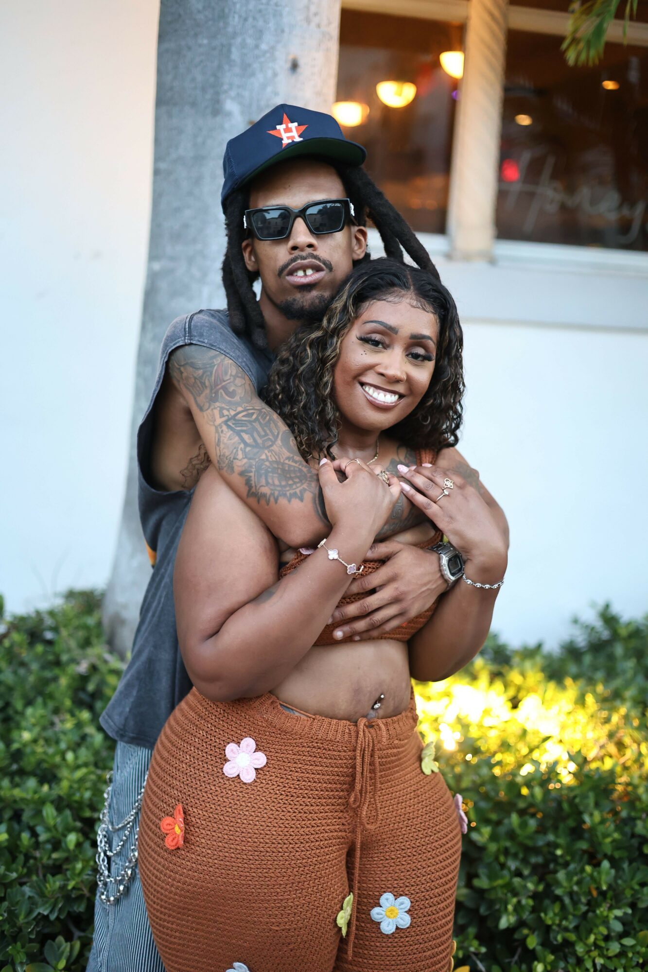 A smiling woman with curly hair and a man with sunglasses and a cap pose outdoors, embracing each other.
