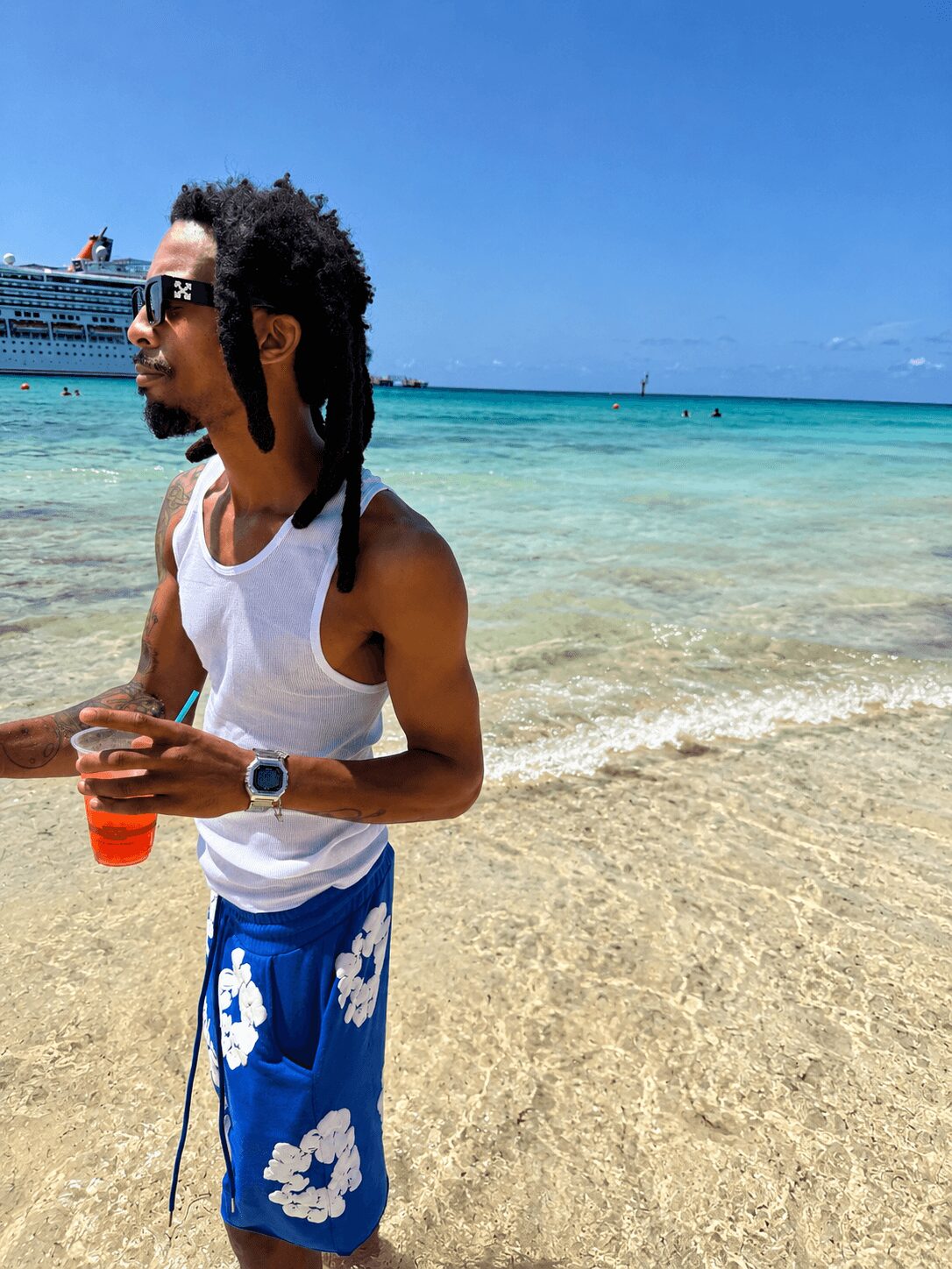 Man with sunglasses and dreadlocks standing on a beach holding a drink, ocean and cruise ship in background.