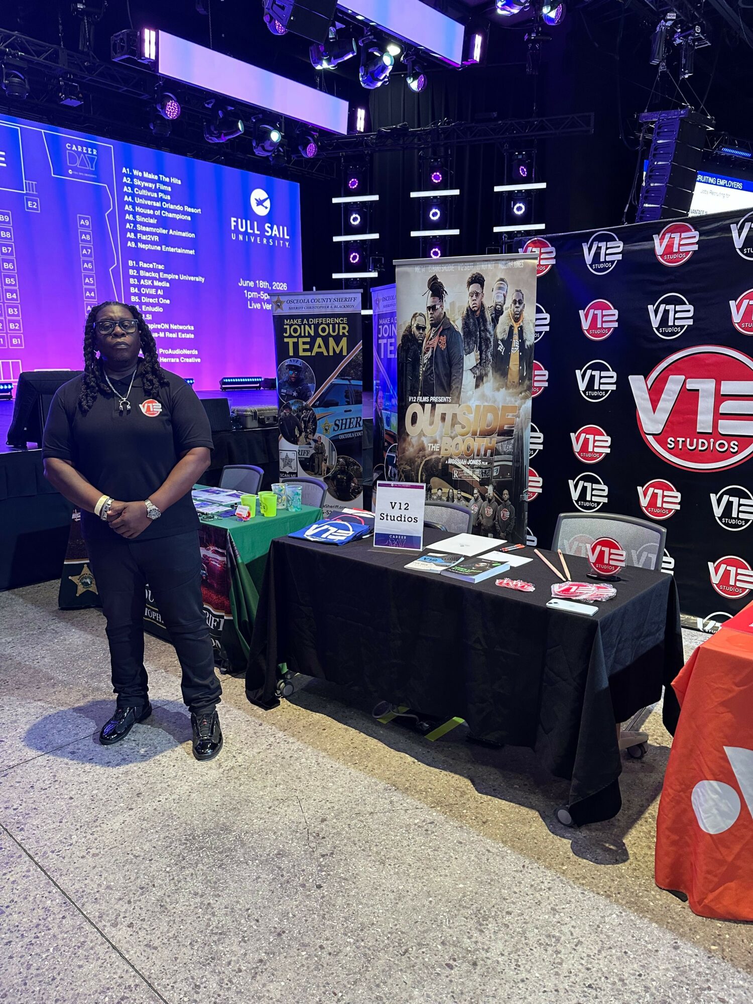 Person standing next to a display table with promotional items and posters, in an indoor event space with blue lighting.