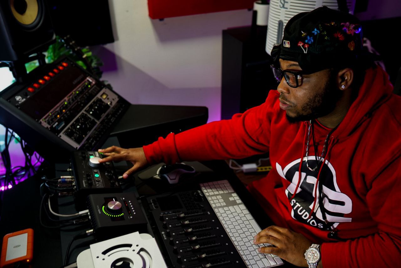 Person wearing glasses and a red hoodie working with audio equipment in a studio, with colorful lights and electronic devices.