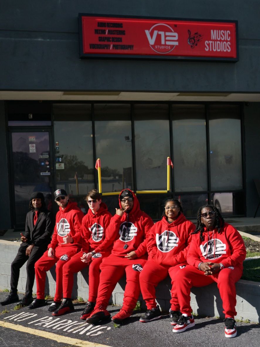 Six people sitting on a bench outside a building with glass windows, wearing red outfits and sunglasses.
