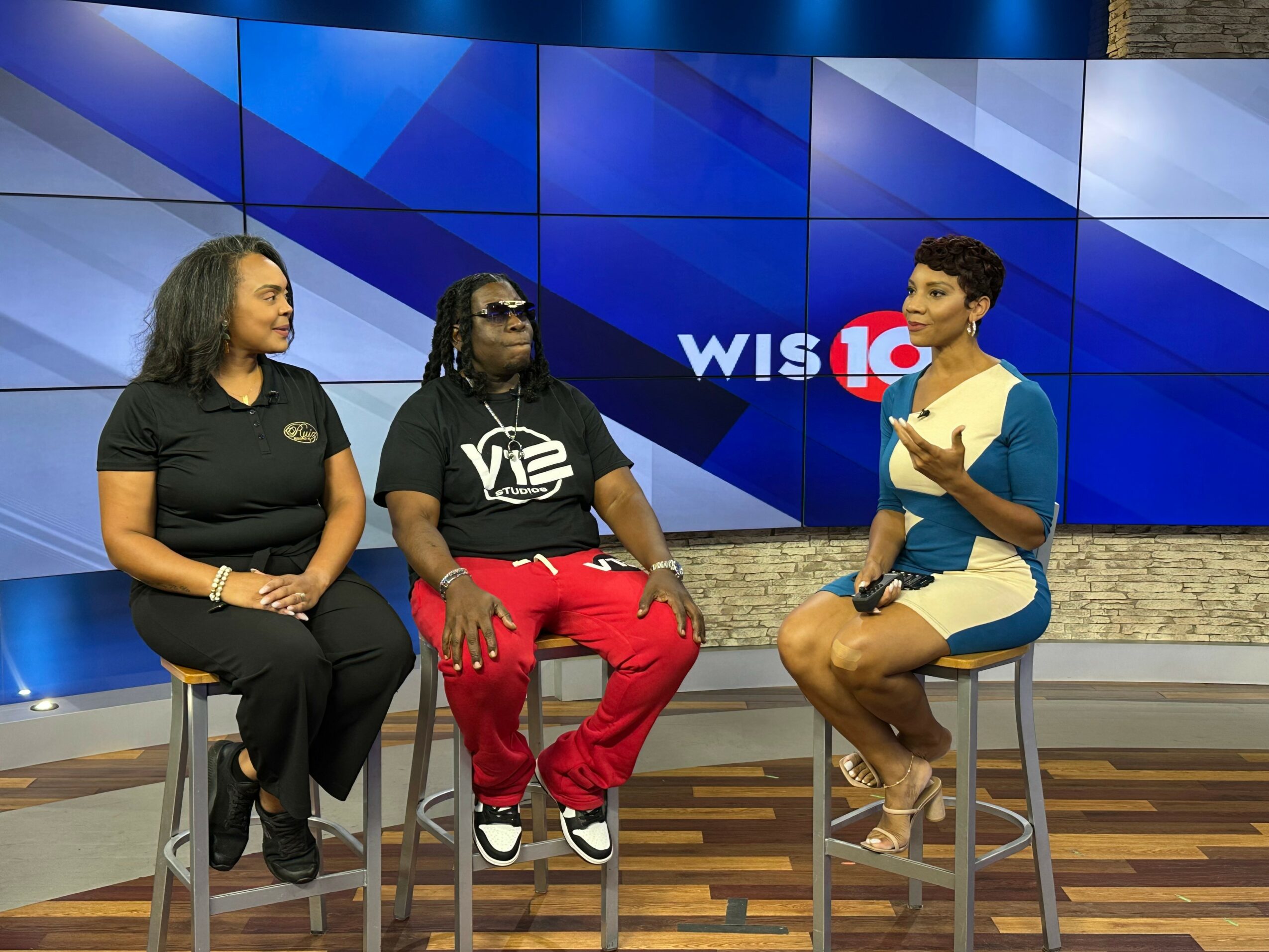 Three women sit on stools in a TV studio with a blue background and a large screen displaying 'WIS 10'.