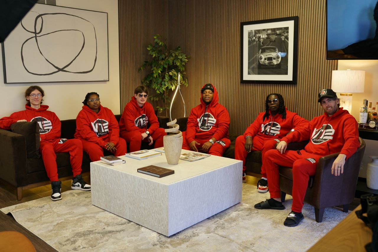 Seven people sit on couches in a room, wearing red hoodies and pants, with a white coffee table in front of them.