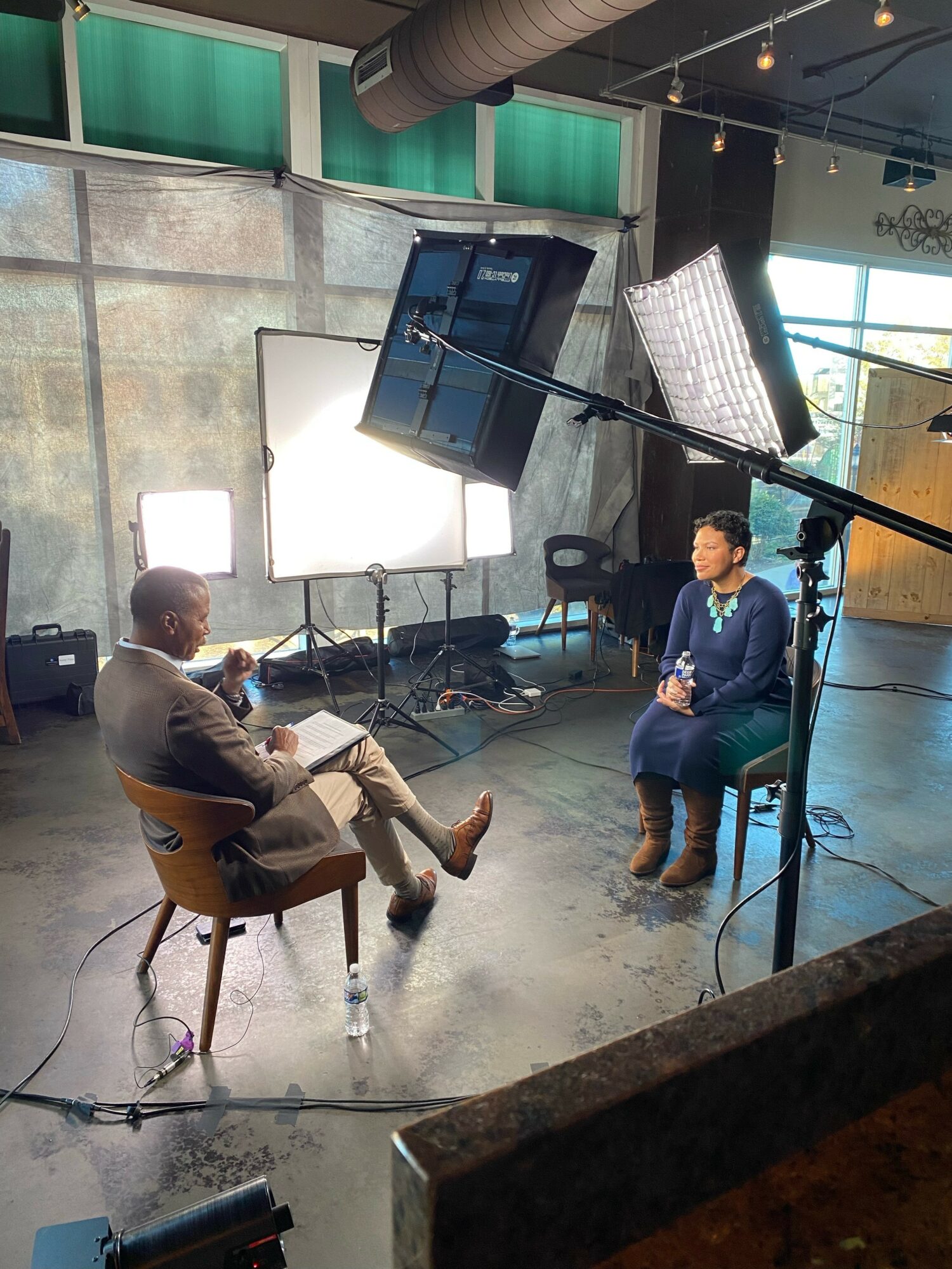 Two people sit in a studio with lighting and camera equipment, engaged in conversation, with large windows in the background.