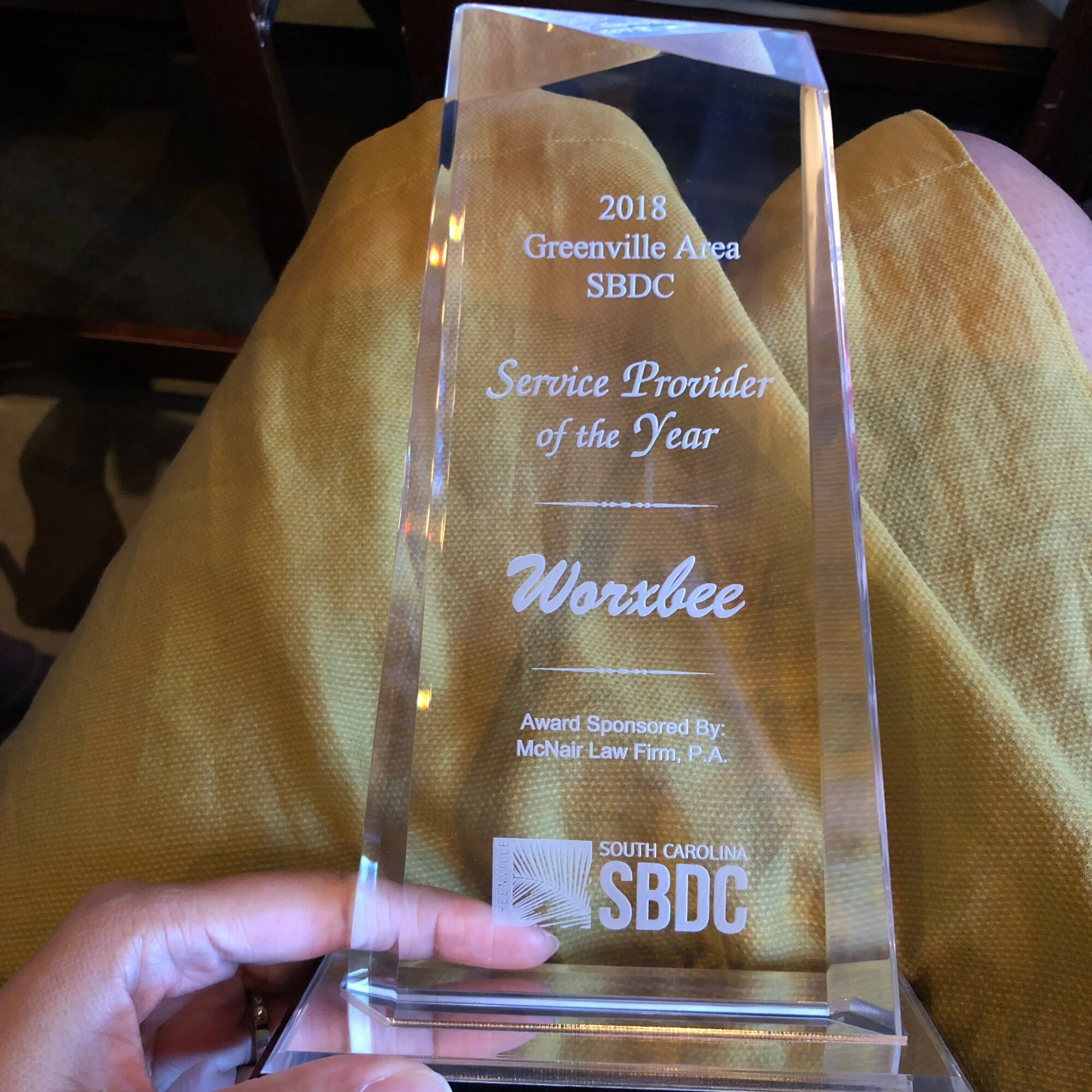 Clear glass award with engraved text, held by a person's hand, resting on person's lap in yellow pants.
