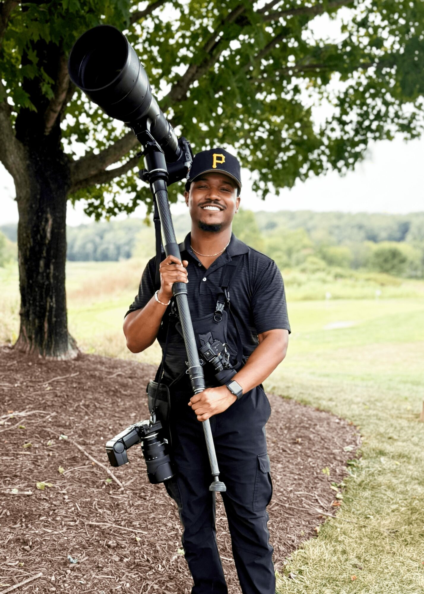 Man outdoors holding a large camera with a long lens, smiling, wearing a black cap with a yellow 'P', black shirt, and pants.