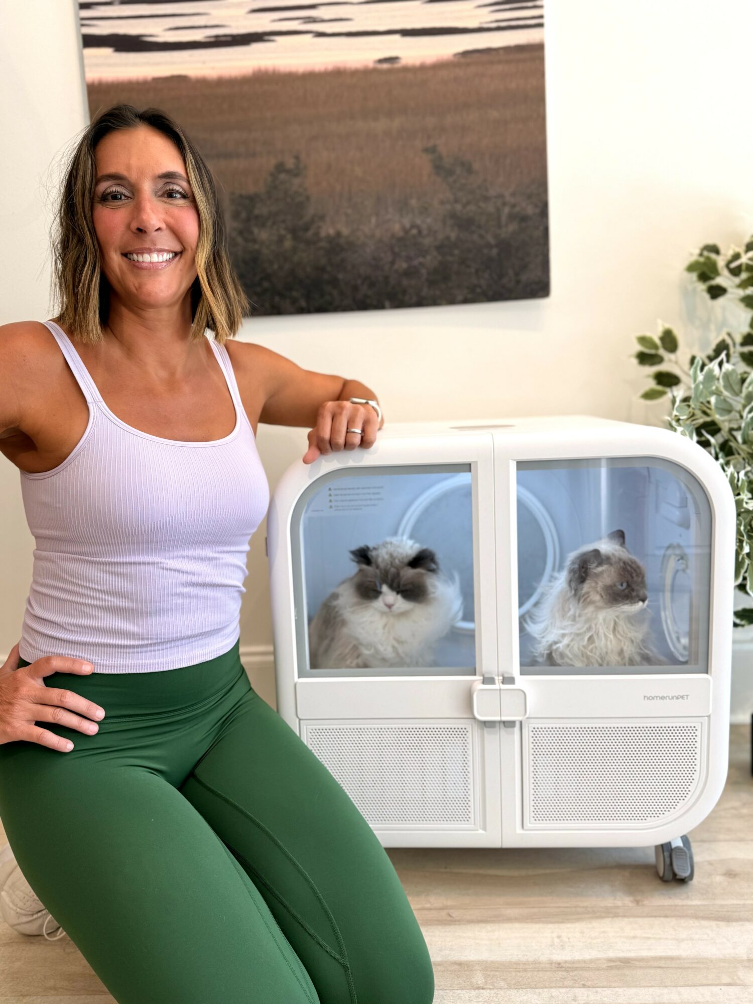 Woman smiling with two cats in a white enclosure, standing indoors near a plant and a painting.