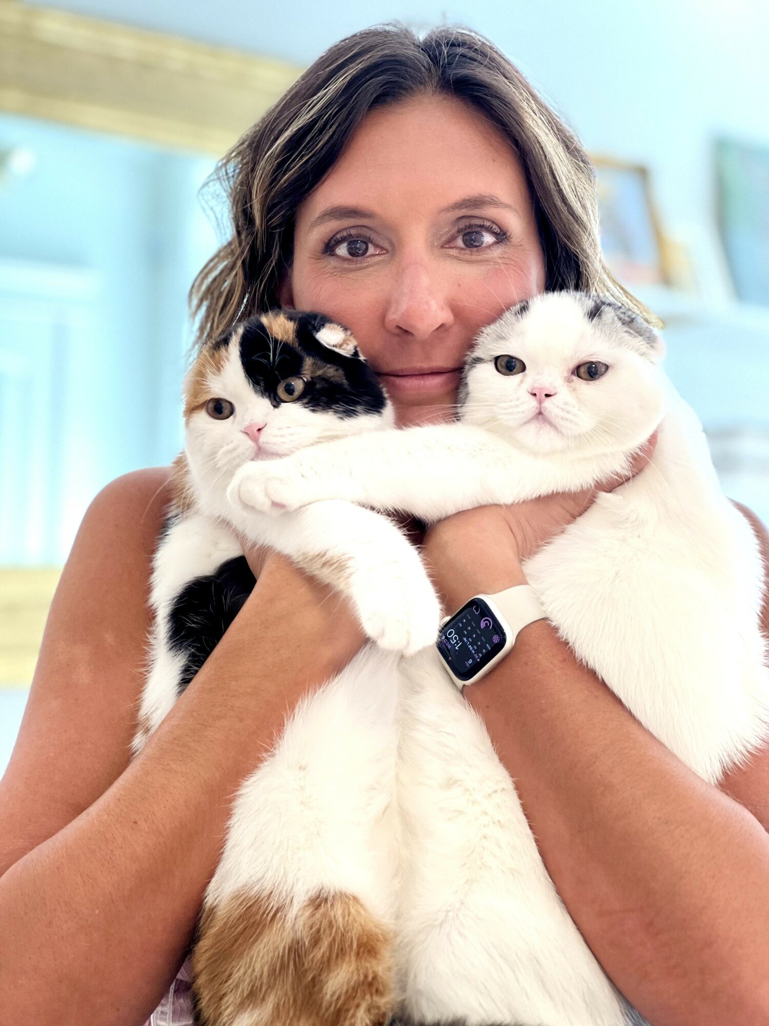 Woman holding two cats close to her face indoors, smiling, with a light blue background.