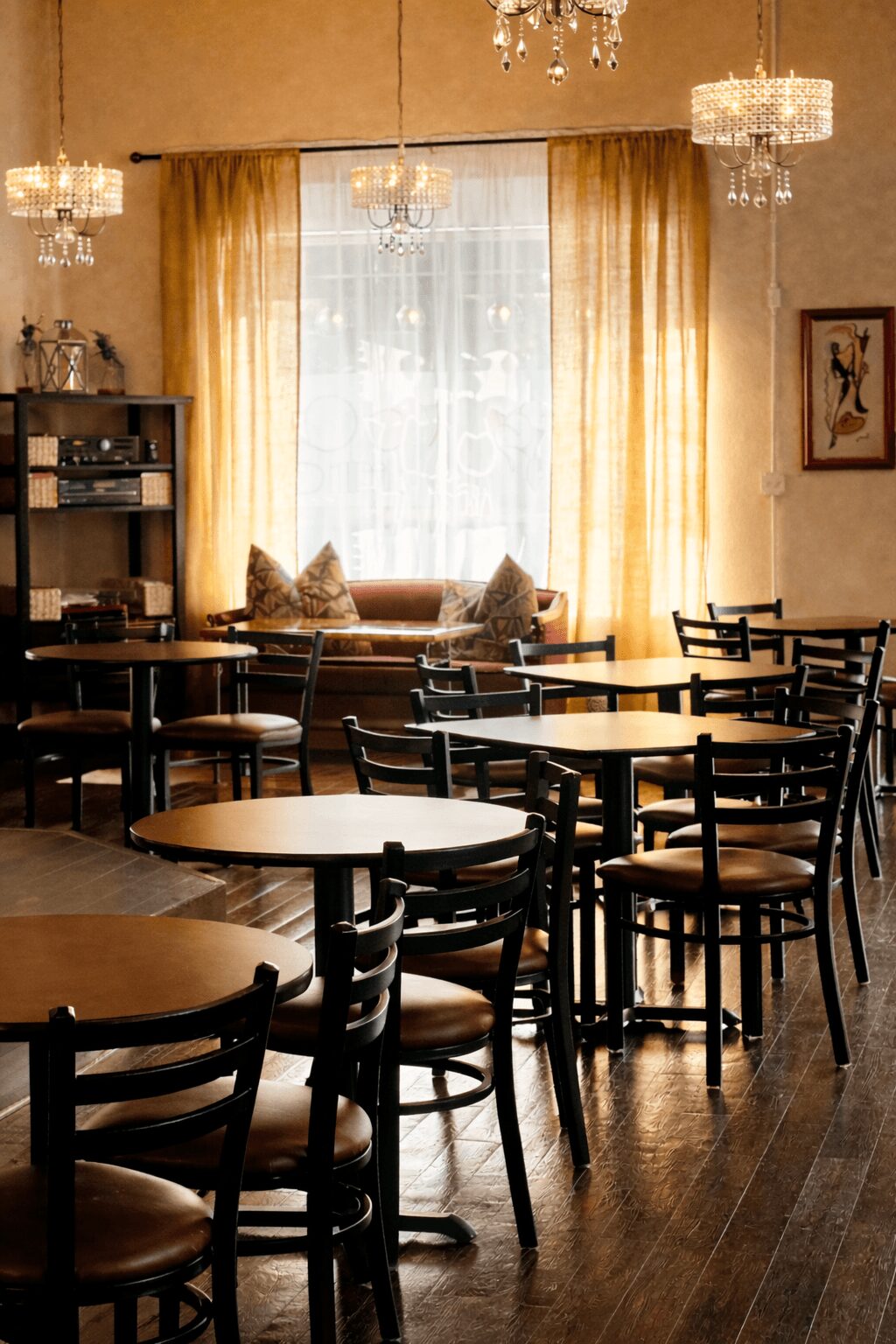 Interior of a cozy room with tables, chairs, a window with curtains, and a sofa with cushions, warm lighting.