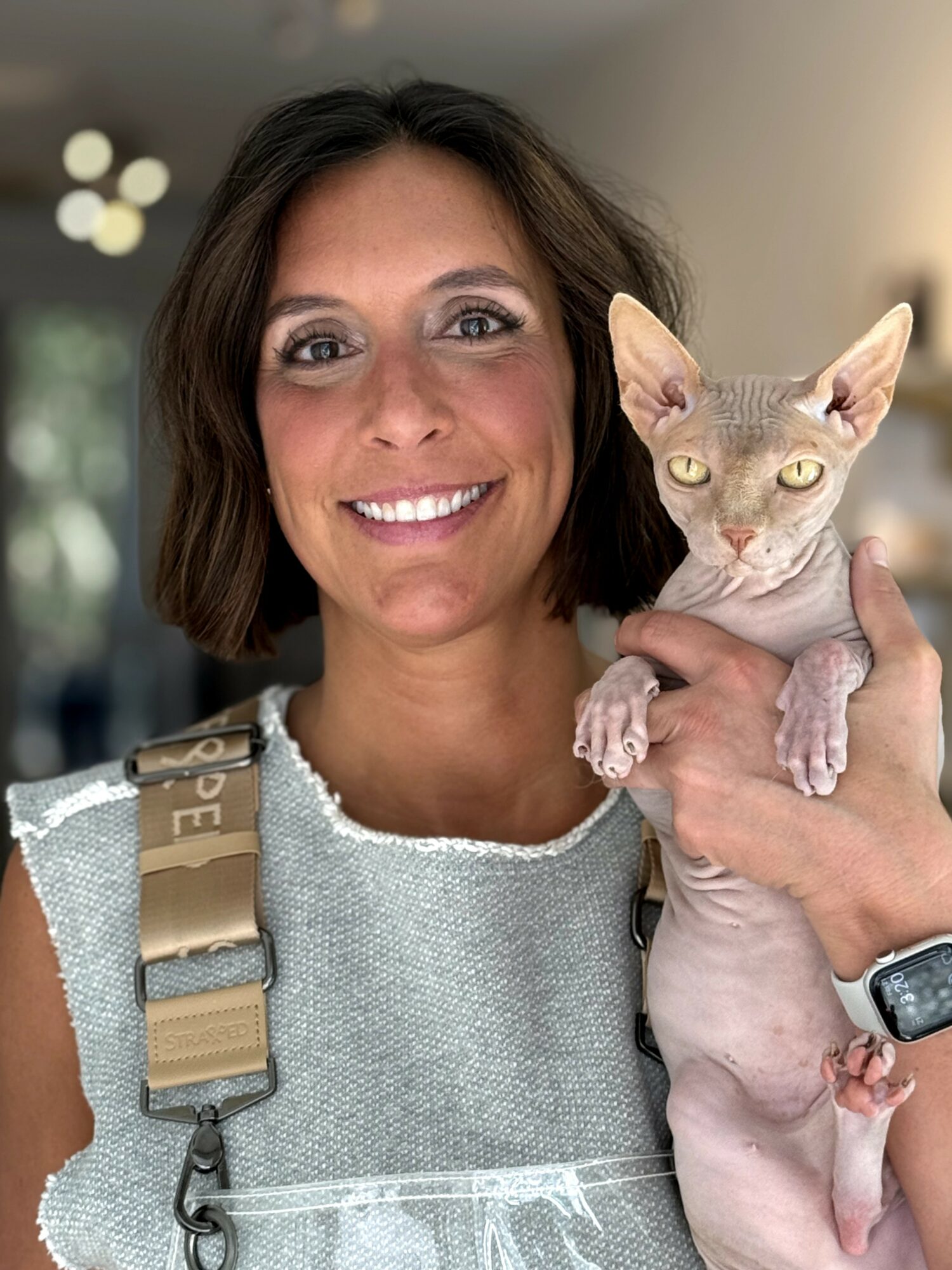 Woman smiling holding a hairless cat indoors, background with blurred lights, wearing a light-colored top and a watch.