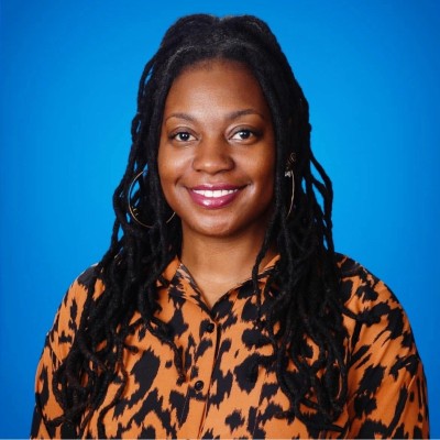 Portrait of a smiling woman with long dreadlocks wearing a patterned shirt against a blue background.