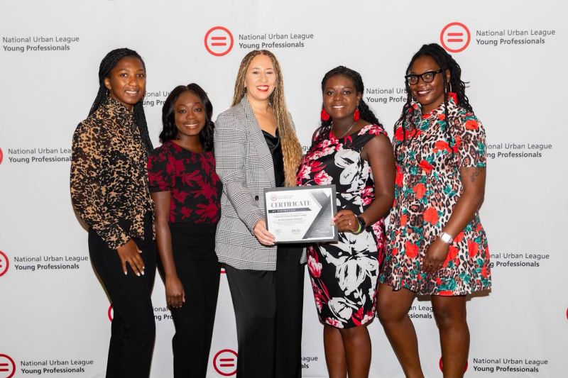 Five women standing together, one holding a certificate, smiling in front of a white background with text. Word count: 24