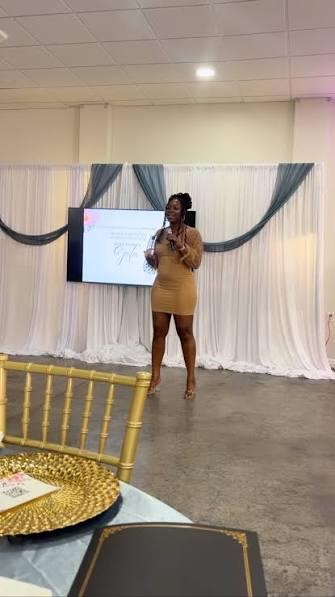 Woman standing on stage giving a presentation, with a screen and draped backdrop behind her.