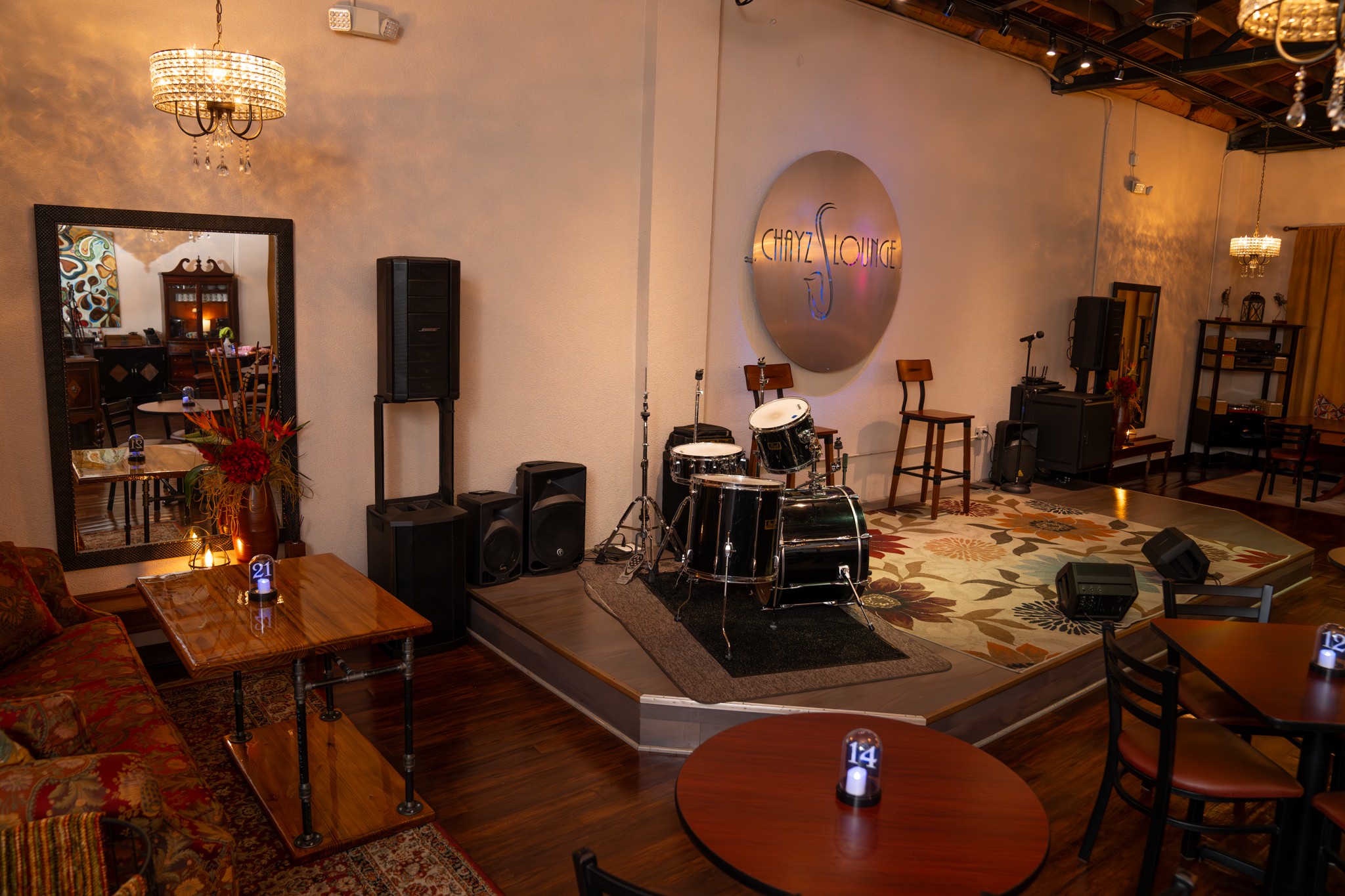 Stage with drums, speakers, and chairs in a cozy, dimly lit venue with wooden floors and decorative lighting.