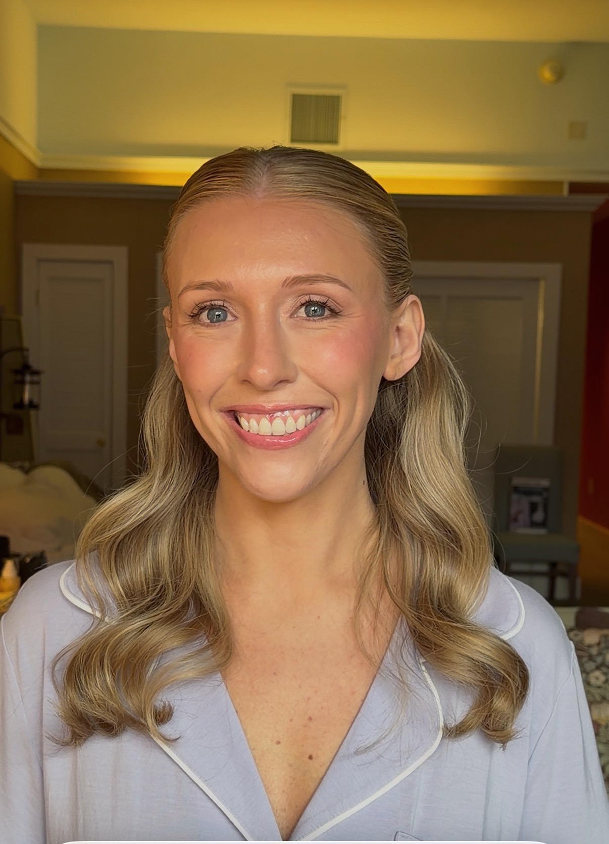 Smiling woman with wavy blonde hair in a light-colored top indoors, background with doors and warm lighting.