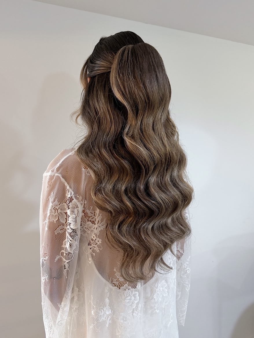 Back view of a woman with long, wavy brown hair wearing a lace dress, standing against a plain wall.