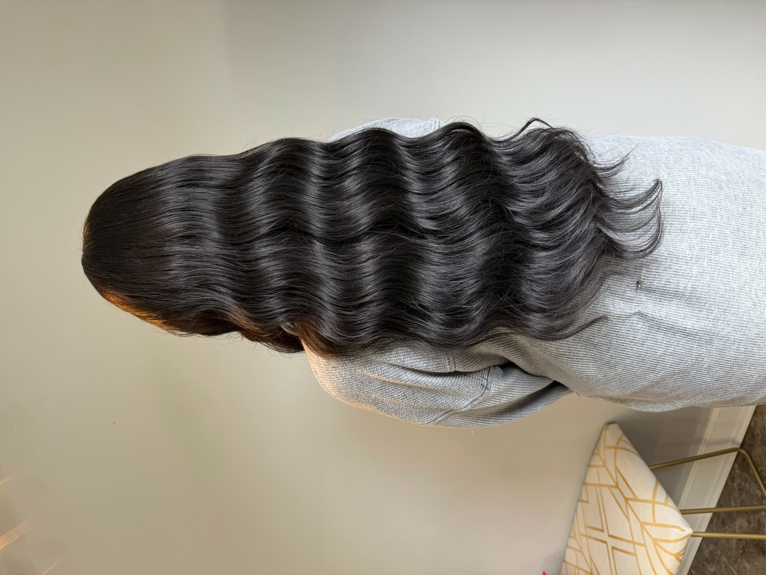 Person with long, wavy, dark hair viewed from the back, wearing a gray top, standing indoors.