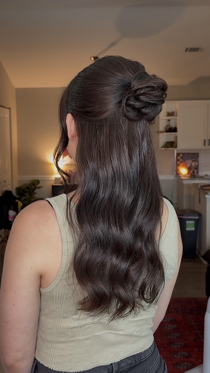Woman with long, wavy dark hair styled in a half-up bun, facing away in a room with kitchen background.