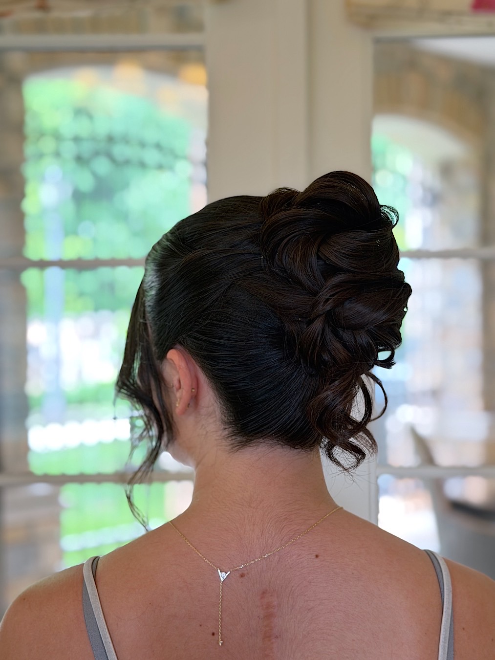 Back view of a woman with an elegant updo hairstyle, wearing a light-colored top, in front of a window with blurred outdoor scenery.