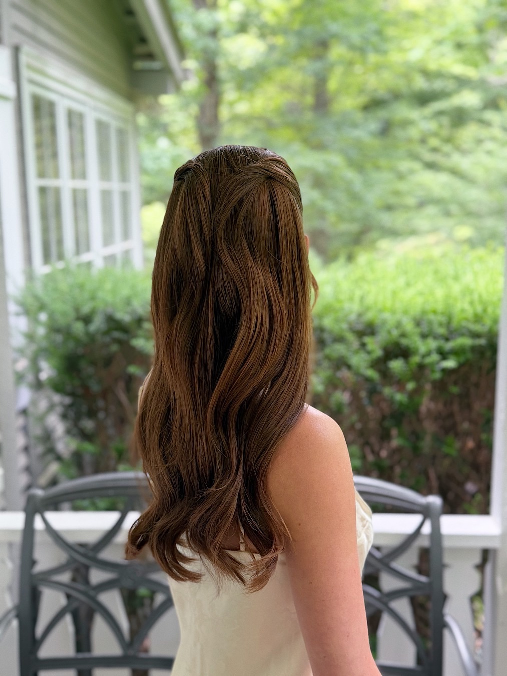 Back view of a woman with long, wavy brown hair standing outdoors near a house with greenery in the background.