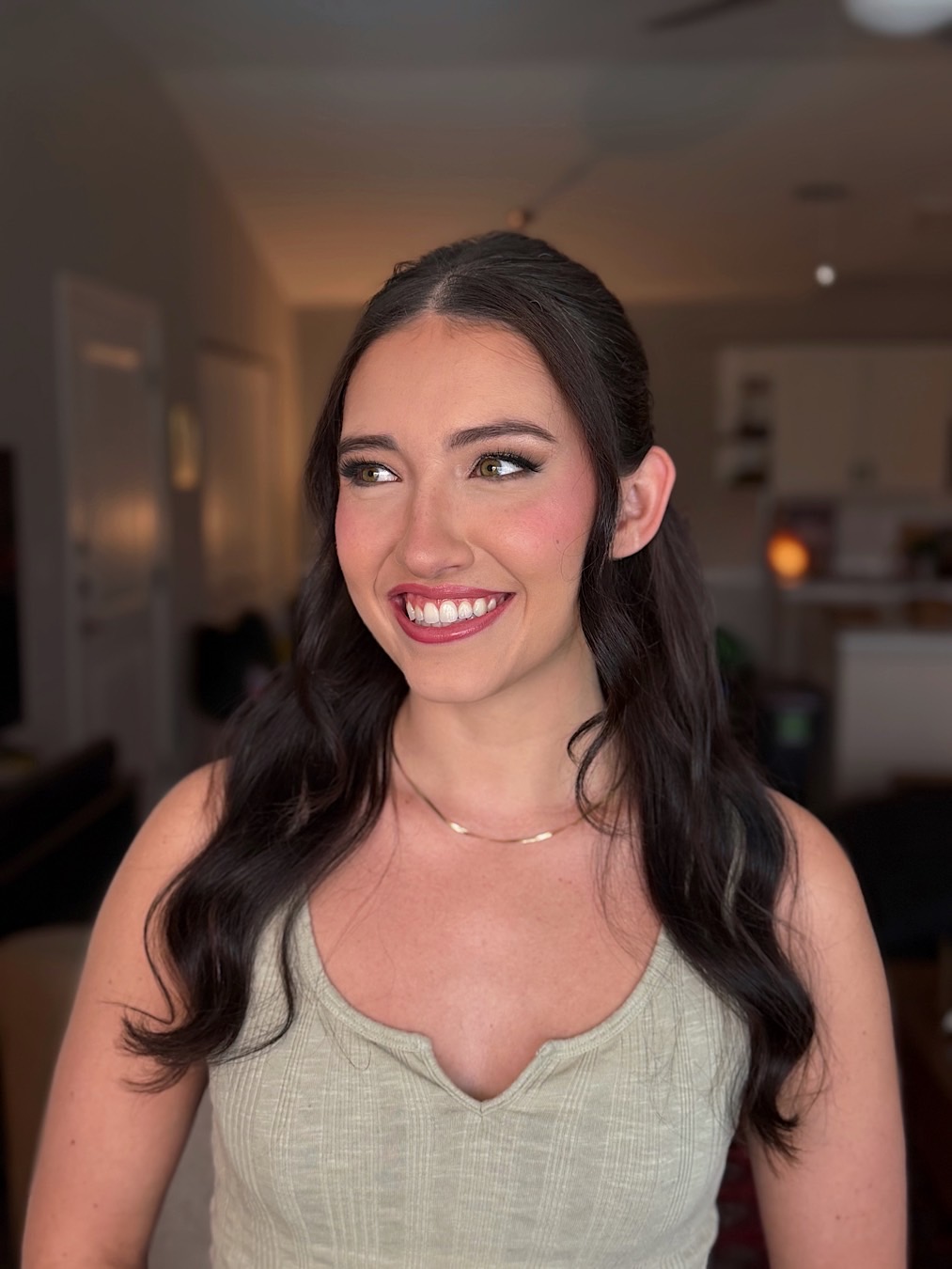 Young woman with long dark hair smiling, wearing a light-colored top, in a home setting.