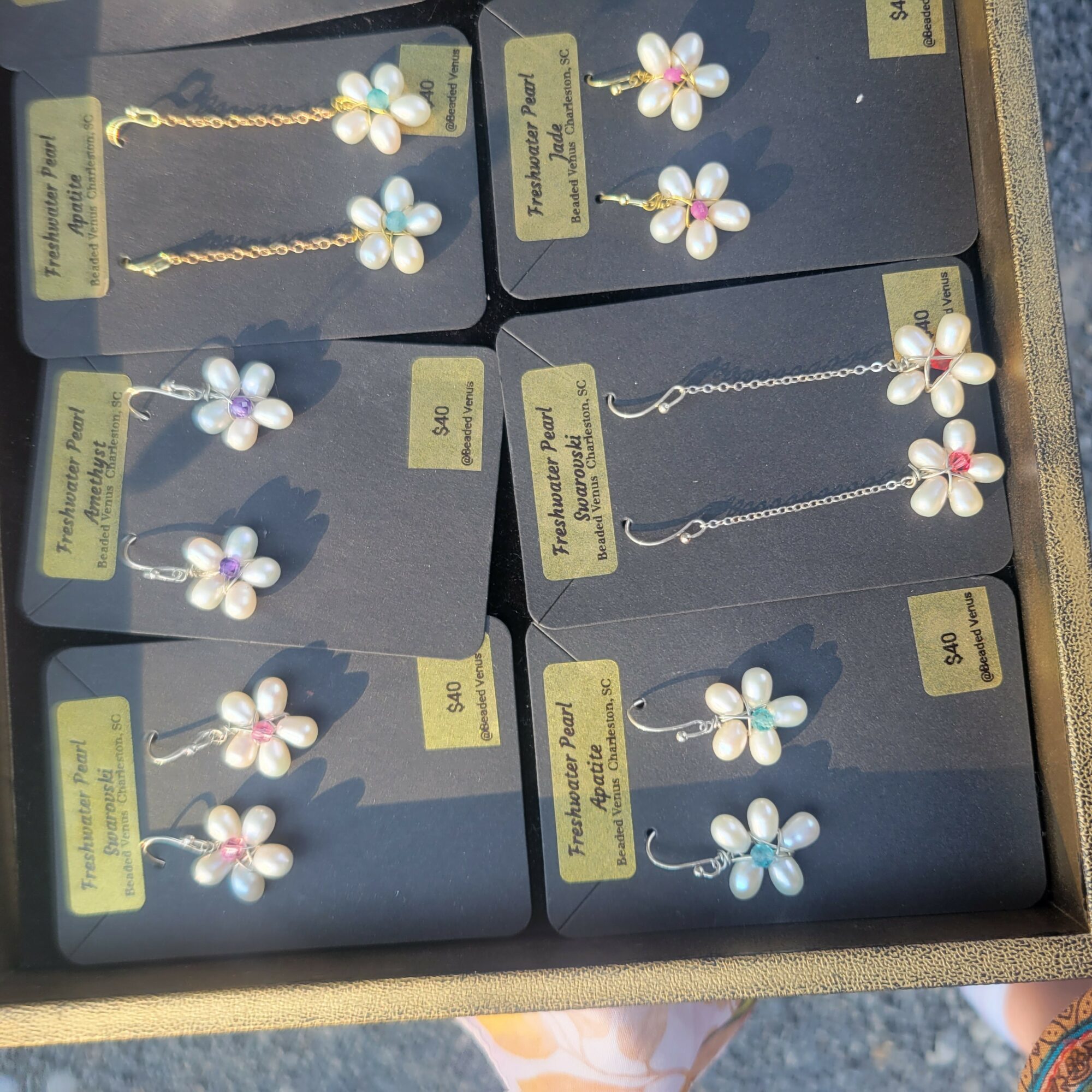 Display of flower-shaped earrings with pearl and pink or blue accents on black cards, arranged in a box.