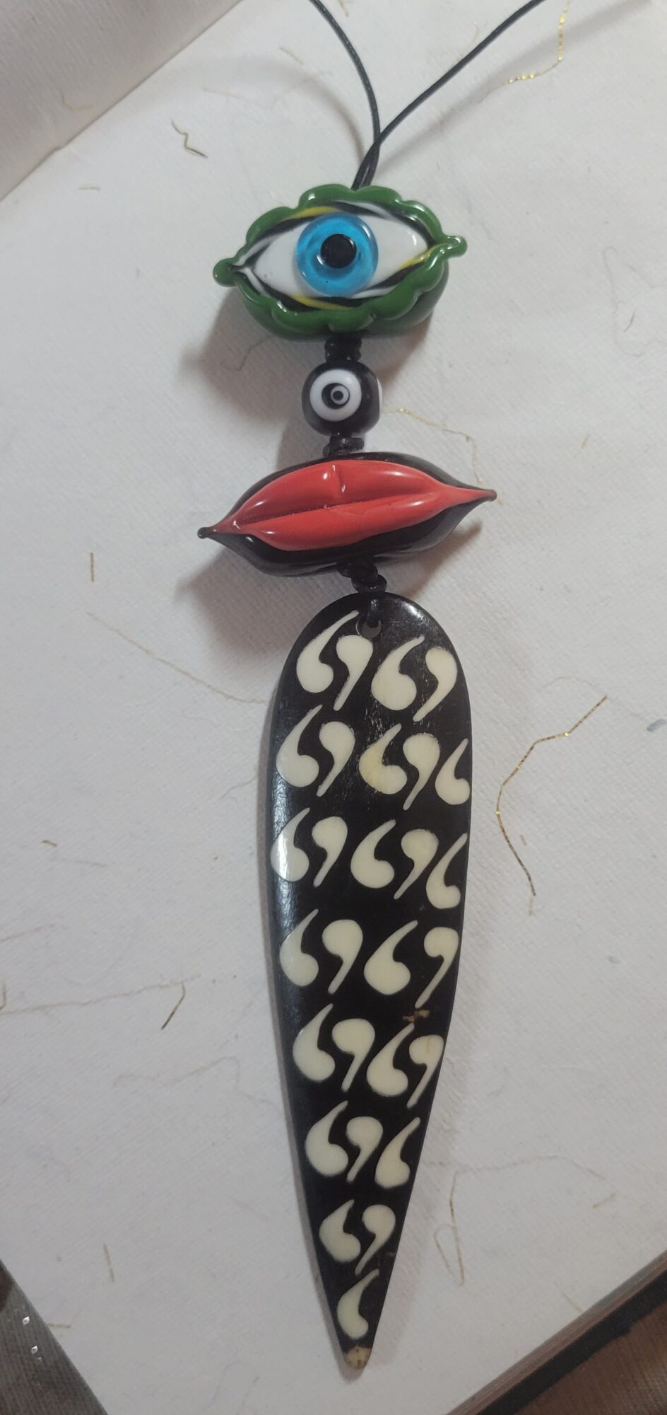 Decorative hanging object with an eye, lips, and a patterned elongated shape, hanging from a string on a white surface.