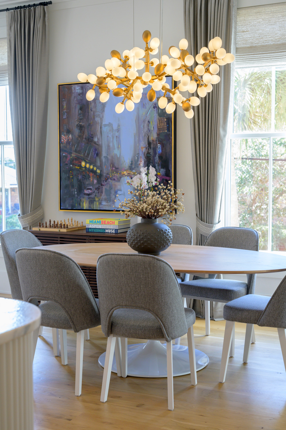 Dining room with a round table, six gray chairs, a large vase with flowers, a chandelier, and a window with curtains.