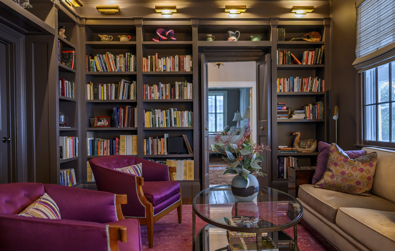 Living room with bookshelves, purple and beige sofas, a glass coffee table with a vase of flowers, and windows with blinds.
