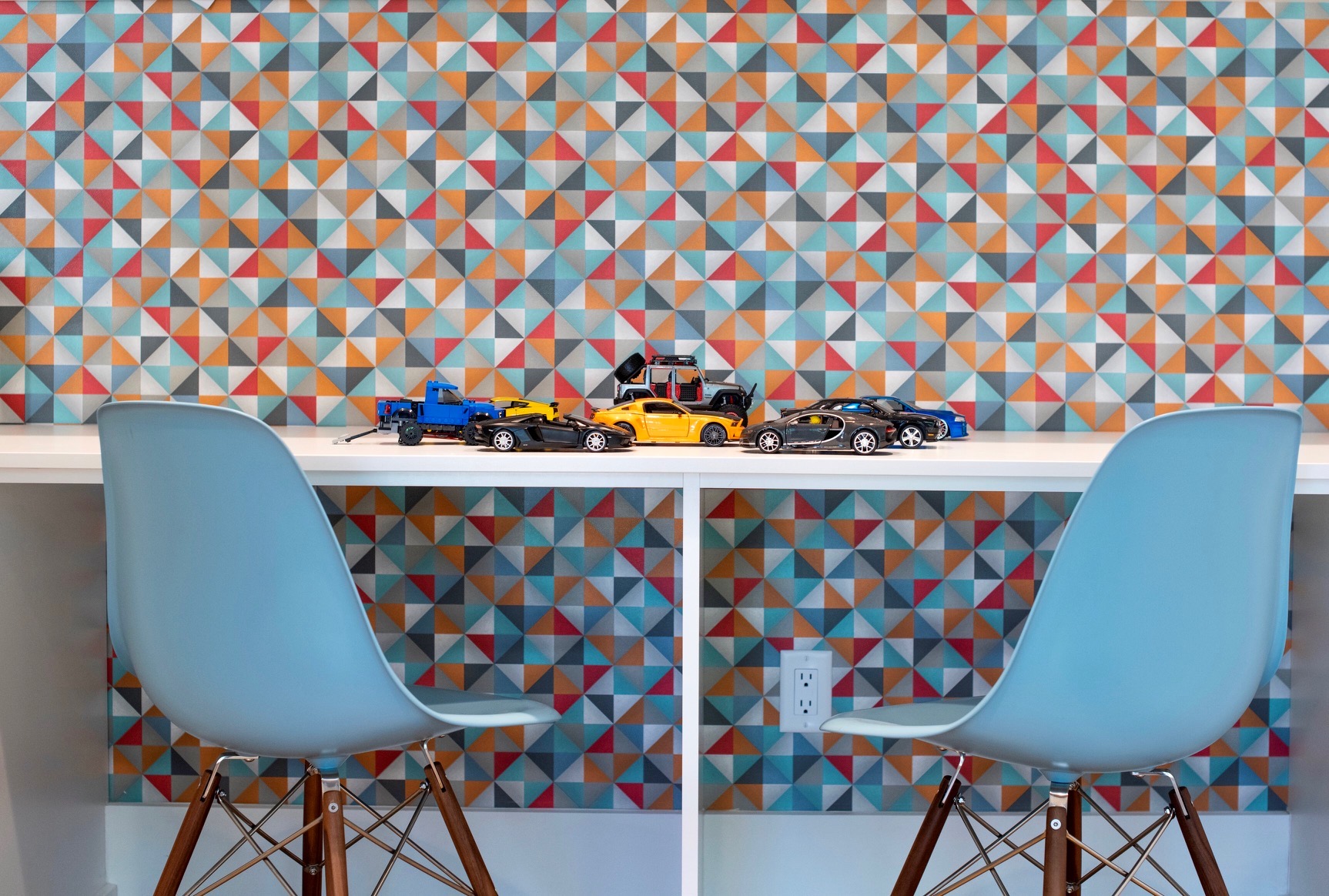 Two blue chairs face a table with toy cars, against a colorful geometric patterned wall.