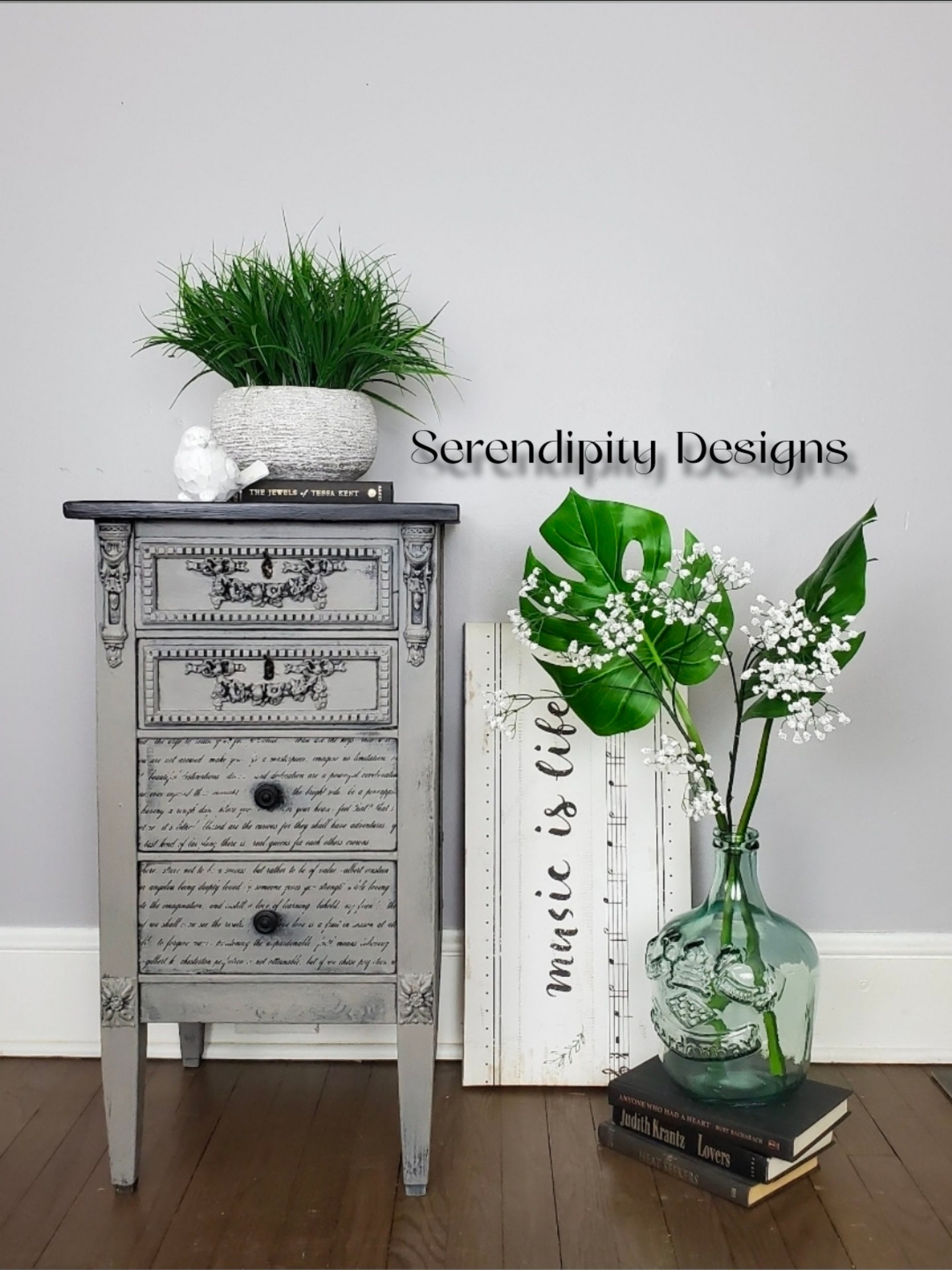 Decorative gray dresser with a potted plant, a glass vase with large leaves and white flowers, and books against a light wall.