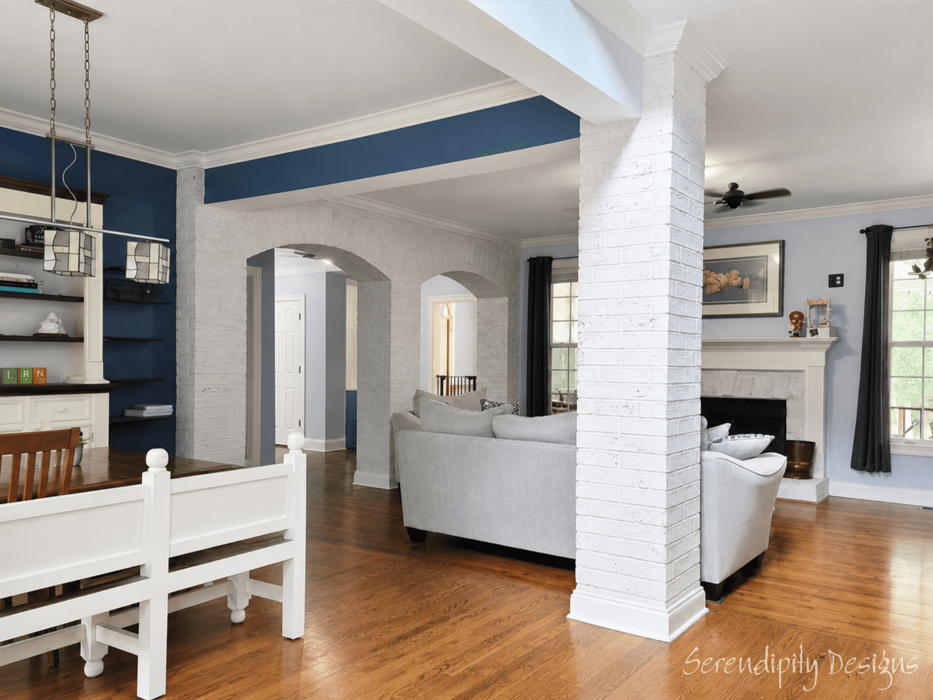 Living room with white brick walls, wooden floor, white sofa, fireplace, and large windows with curtains.
