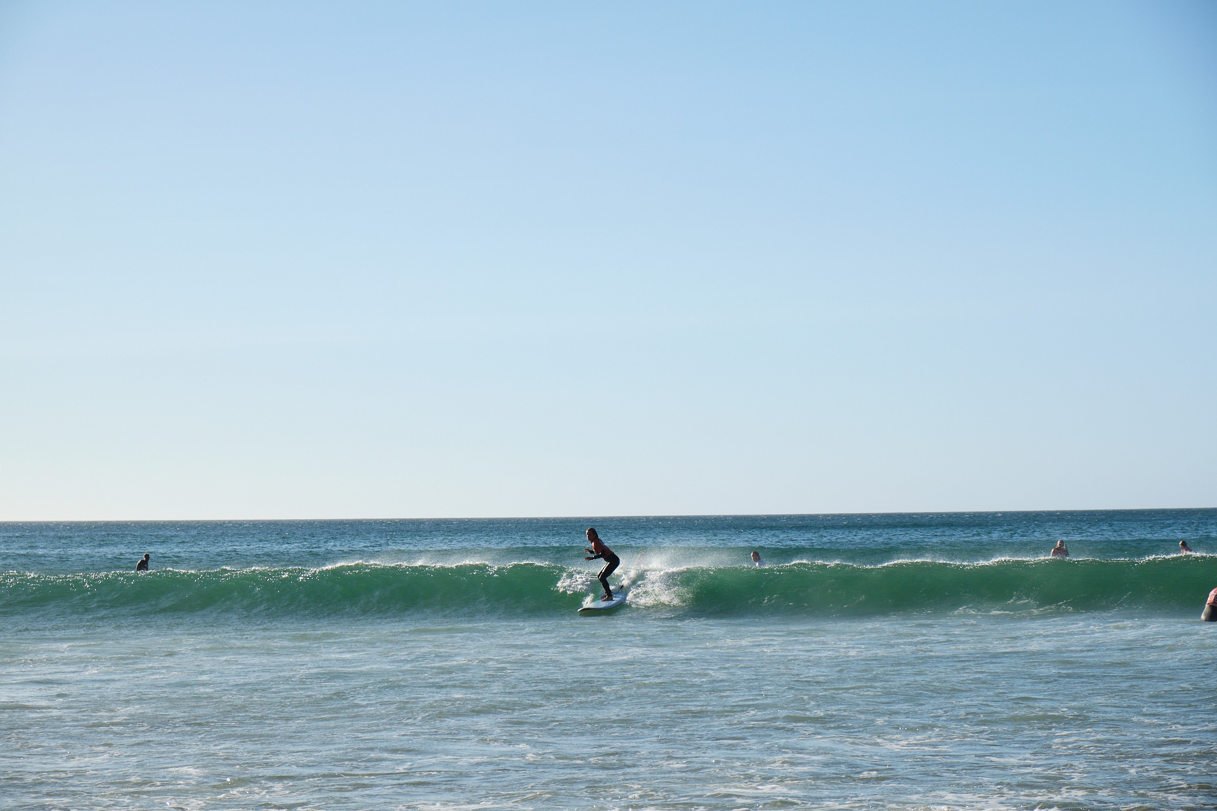 Person surfing on a wave in the ocean with a clear blue sky above, other surfers visible in the water.