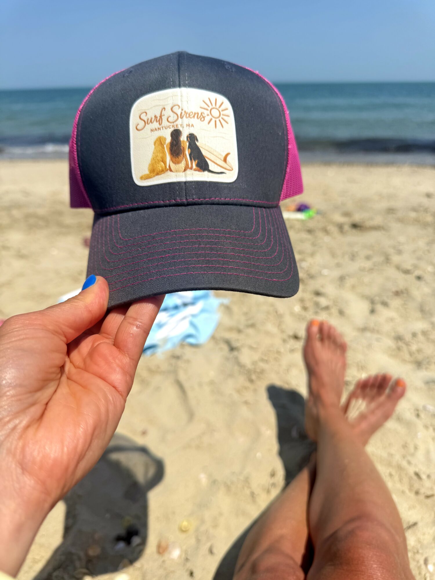 Person relaxing on sandy beach with legs stretched out, holding a cap with a beach scene and text, ocean in background.