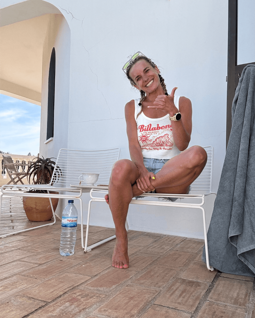 Woman sitting on a white bench, smiling, giving a thumbs-up, with a water bottle on the ground, outdoors.