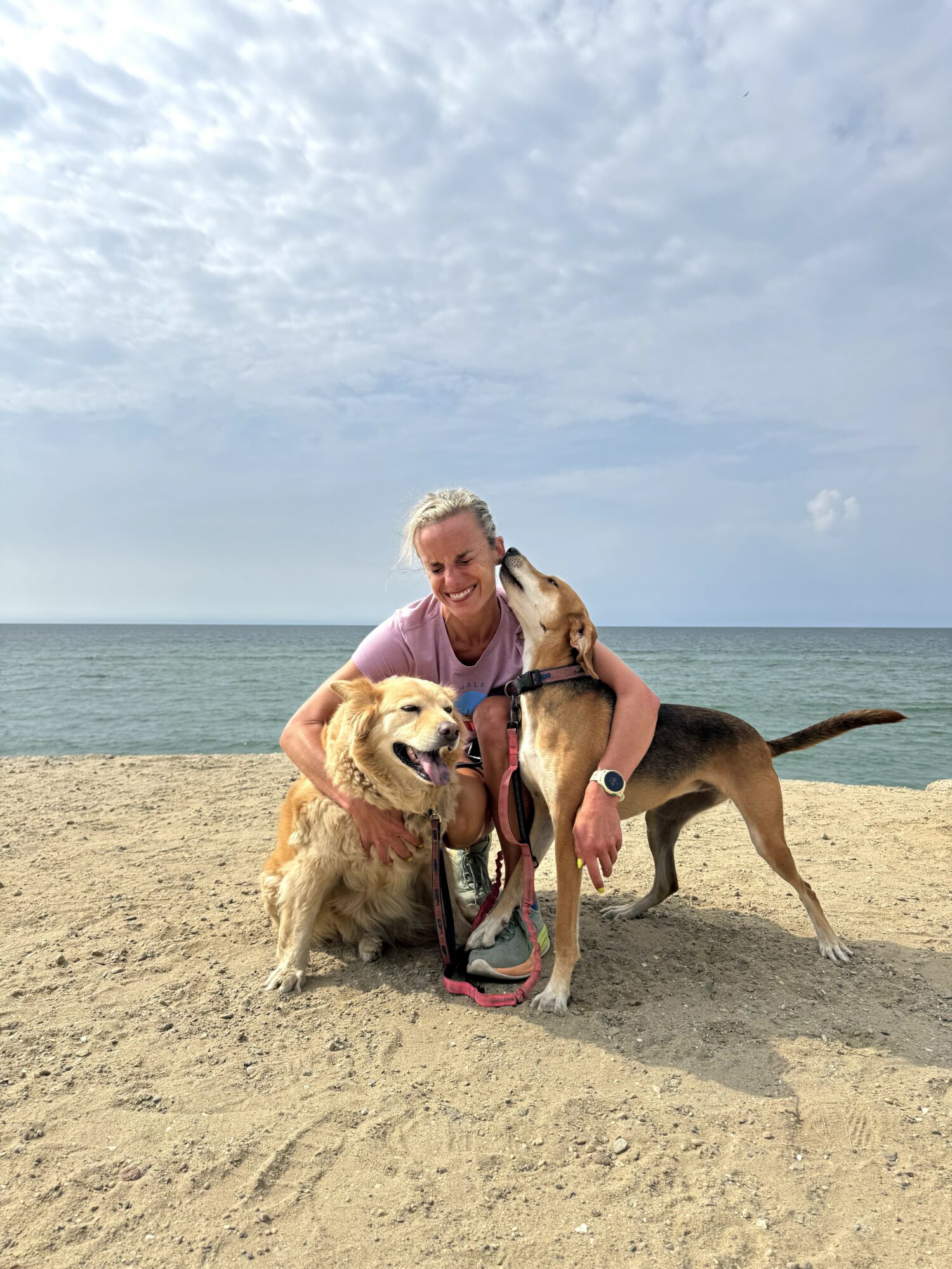 Woman with two dogs on a sandy beach, ocean and sky in background, smiling and holding the dogs' leashes.