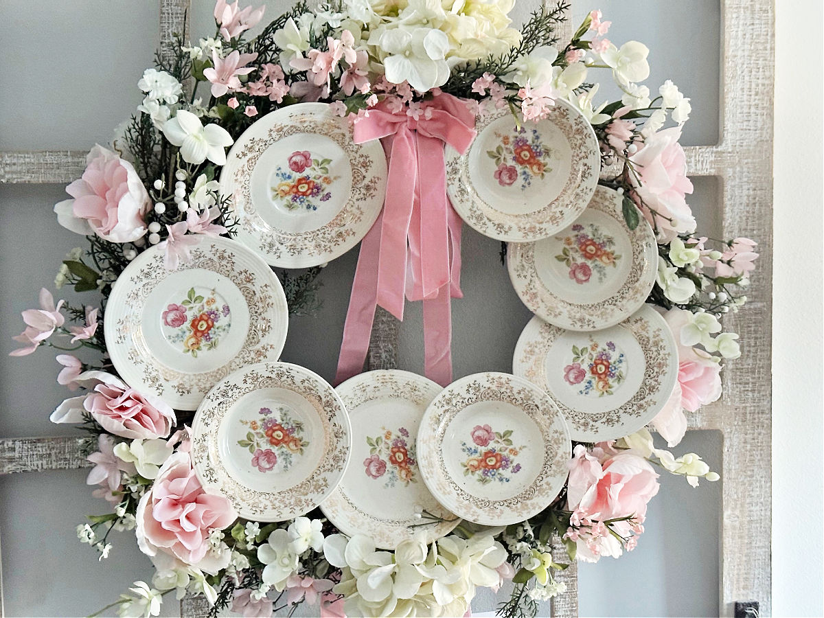 Decorative floral wreath with plates, pink ribbons, and pink and white flowers, hanging on a wall.