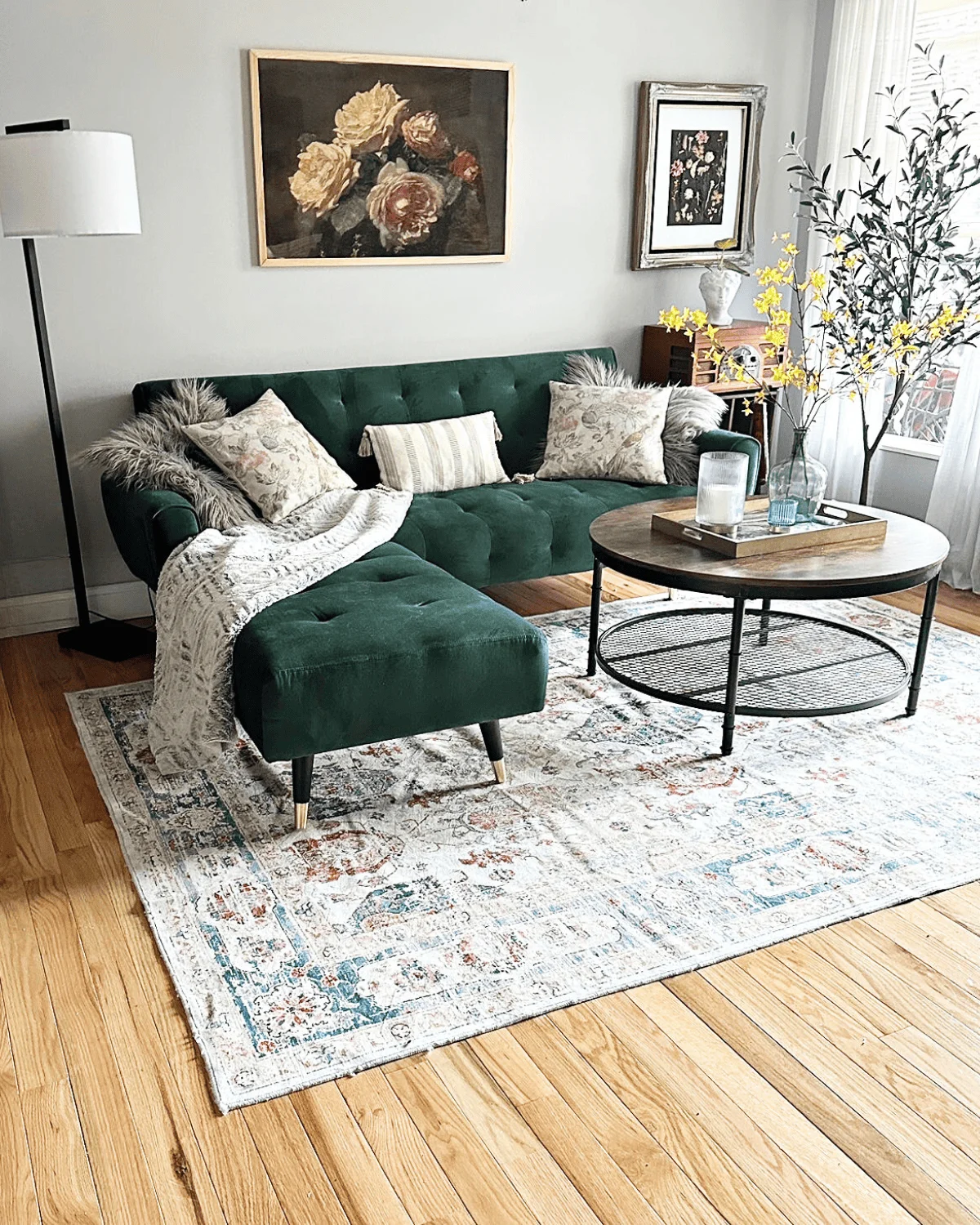 Living room with green velvet sofa, round coffee table, artwork, and a patterned rug on wooden floor.