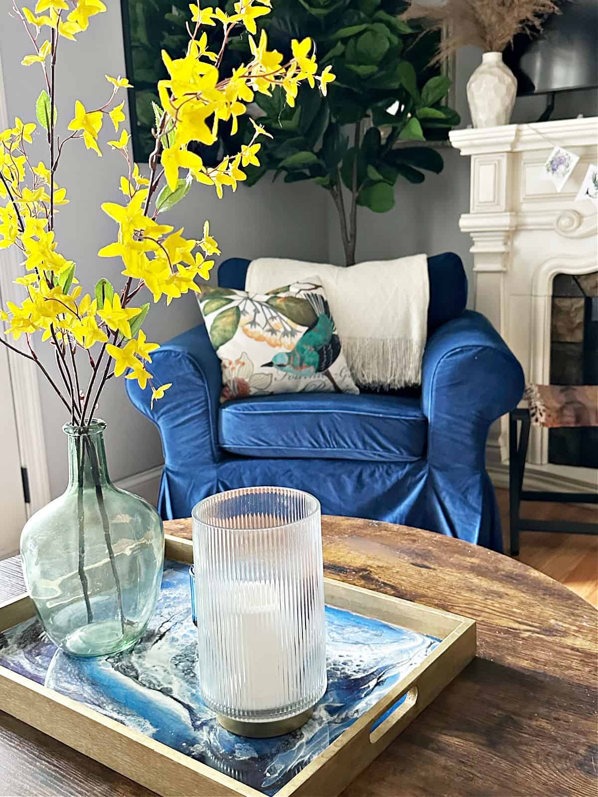 Yellow flowers in a glass vase on a tray, with a blue armchair and decorative pillows in a cozy living room.