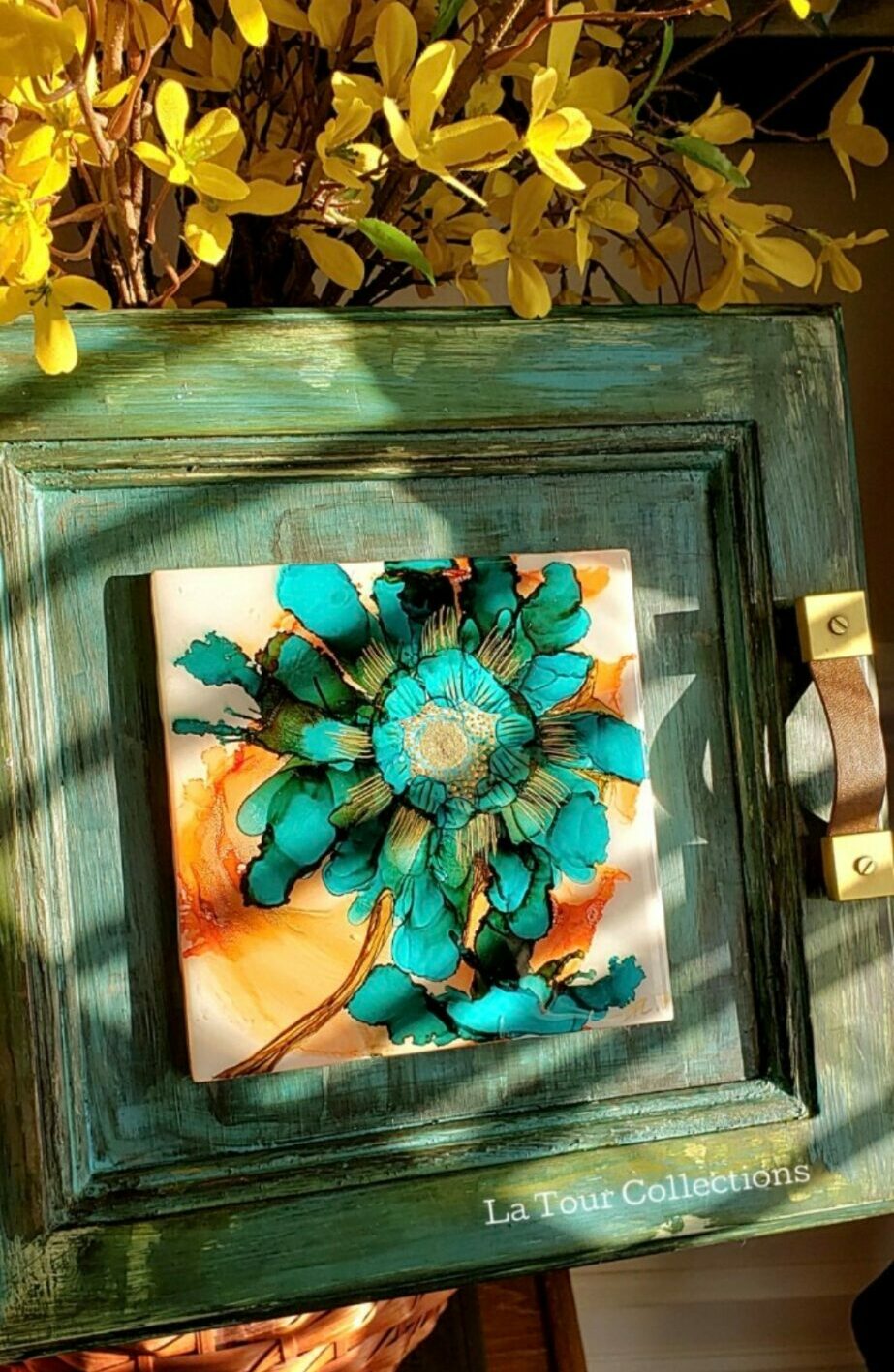Decorative turquoise flower with beads inside a green wooden frame, sunlight casting shadows, background with yellow flowers.