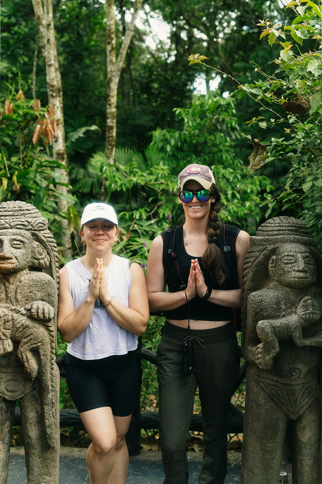 Two women standing between stone statues in a lush green jungle, hands in prayer position, smiling, wearing hats and sunglasses.