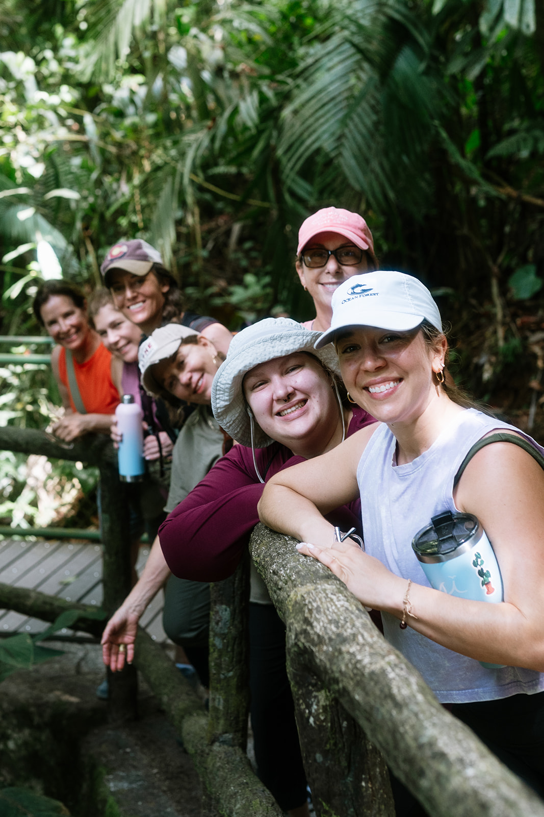 Group of six smiling people leaning on a wooden railing in a lush, green jungle setting.
