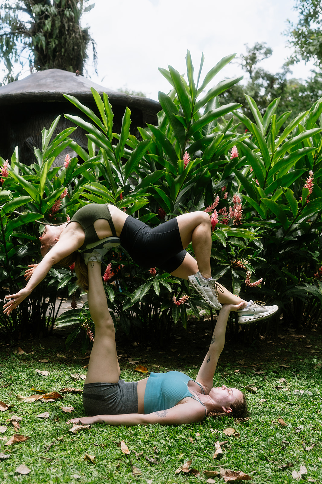 Two people perform an acro yoga pose outdoors with green plants and trees in the background.