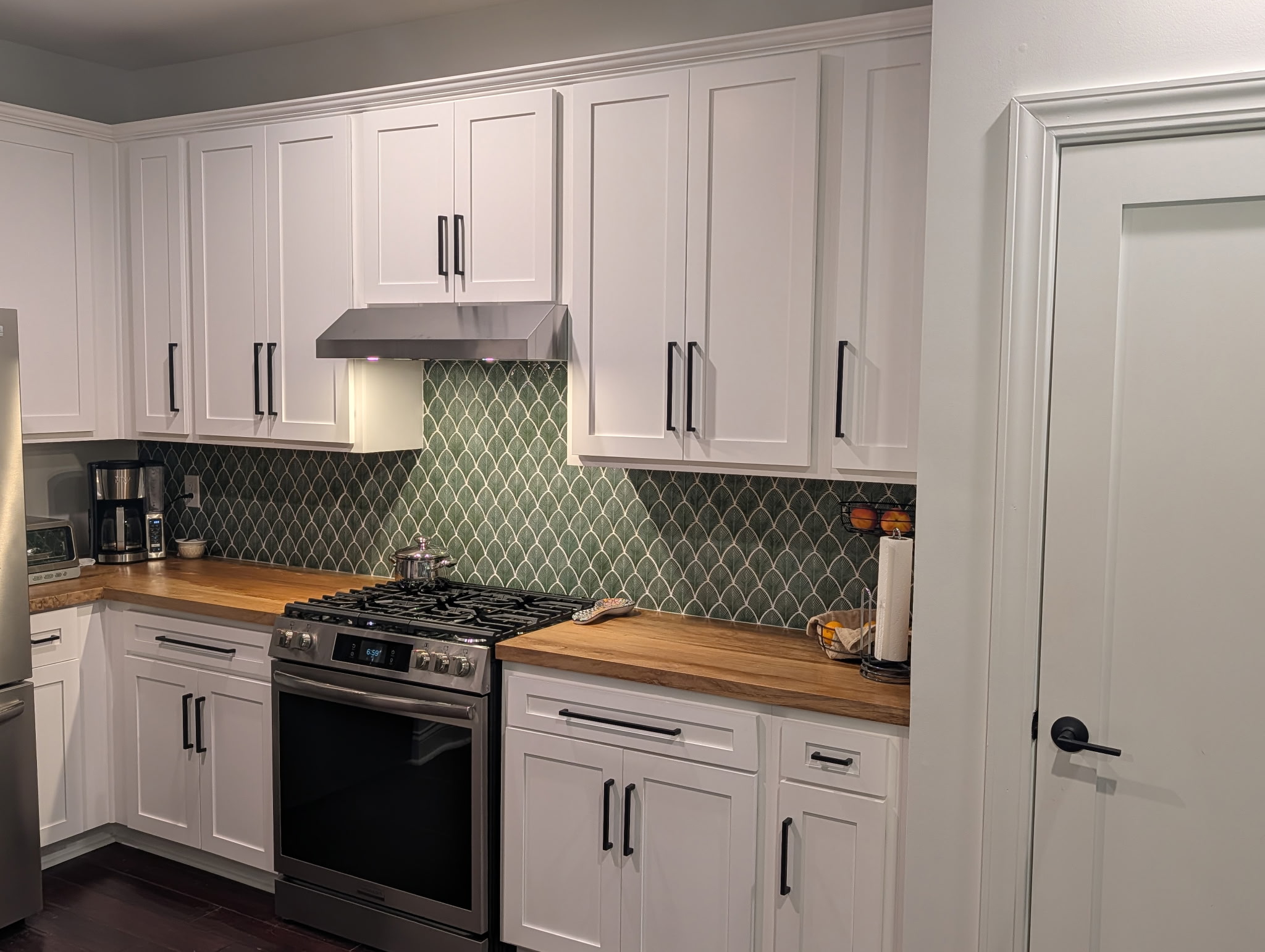 Kitchen with white cabinets, wooden countertop, green tiled backsplash, and a stove with a range hood.