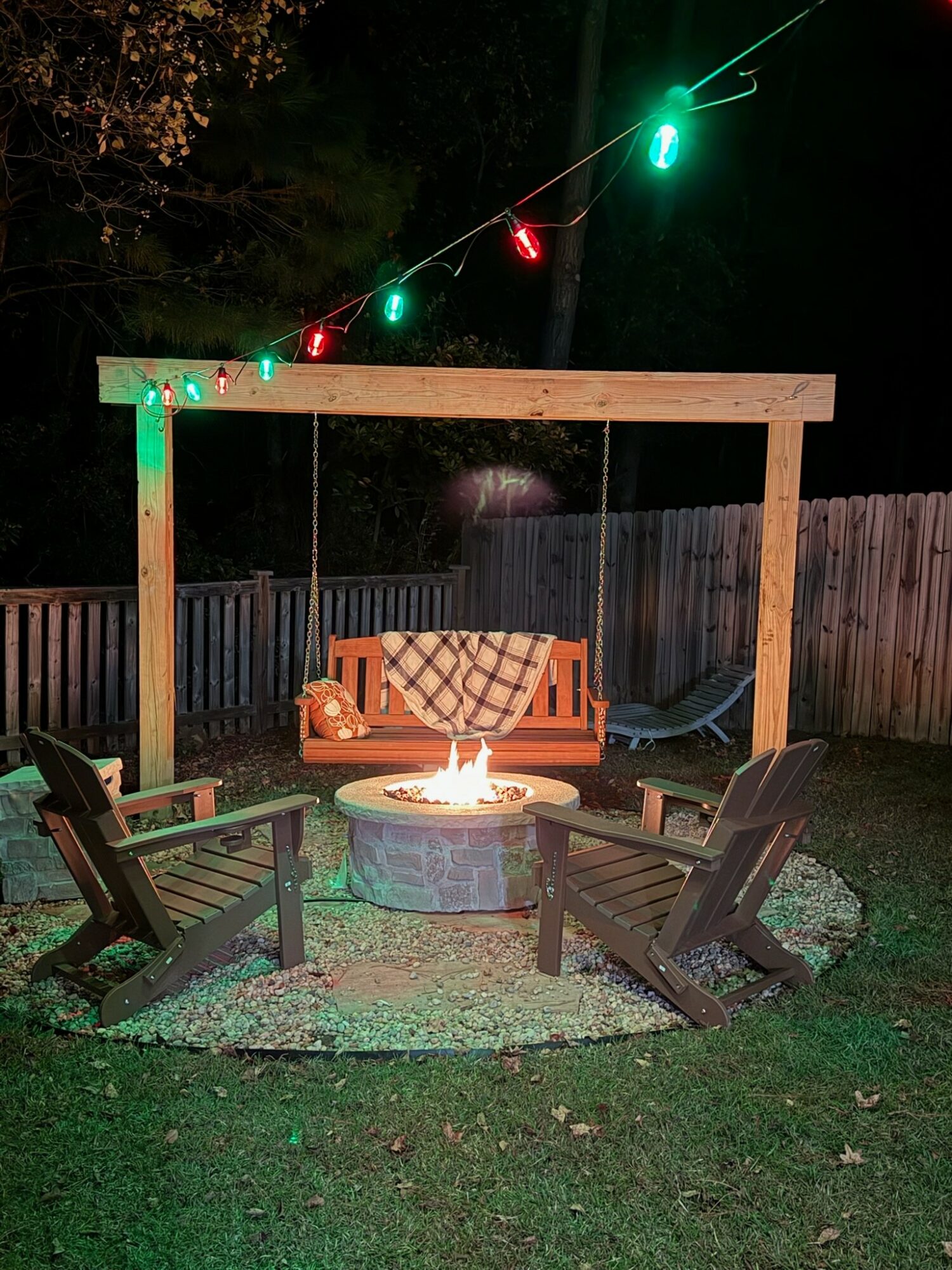 Outdoor backyard scene with a fire pit, four chairs, a wooden pergola, string lights, and a swing, at night.