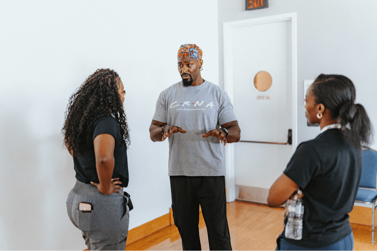 Three people in conversation in a room with white walls and wooden floor, one man and two women, casual attire.
