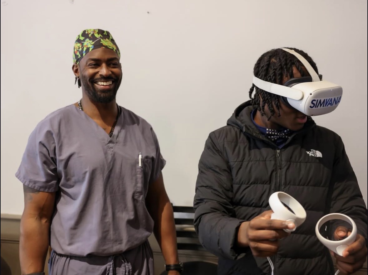 Two men, one smiling and wearing scrubs, the other using a virtual reality headset and holding controllers, stand against a plain background.